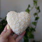 Heart-shaped white soy wax candle with delicate flower textured surface held by a hand against a blurred indoor background