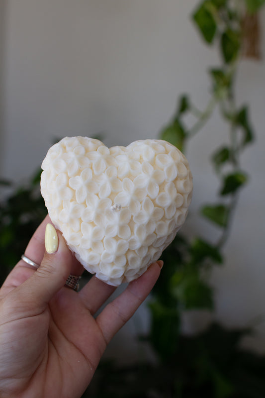 Heart-shaped white soy wax candle with delicate flower textured surface held by a hand against a blurred indoor background