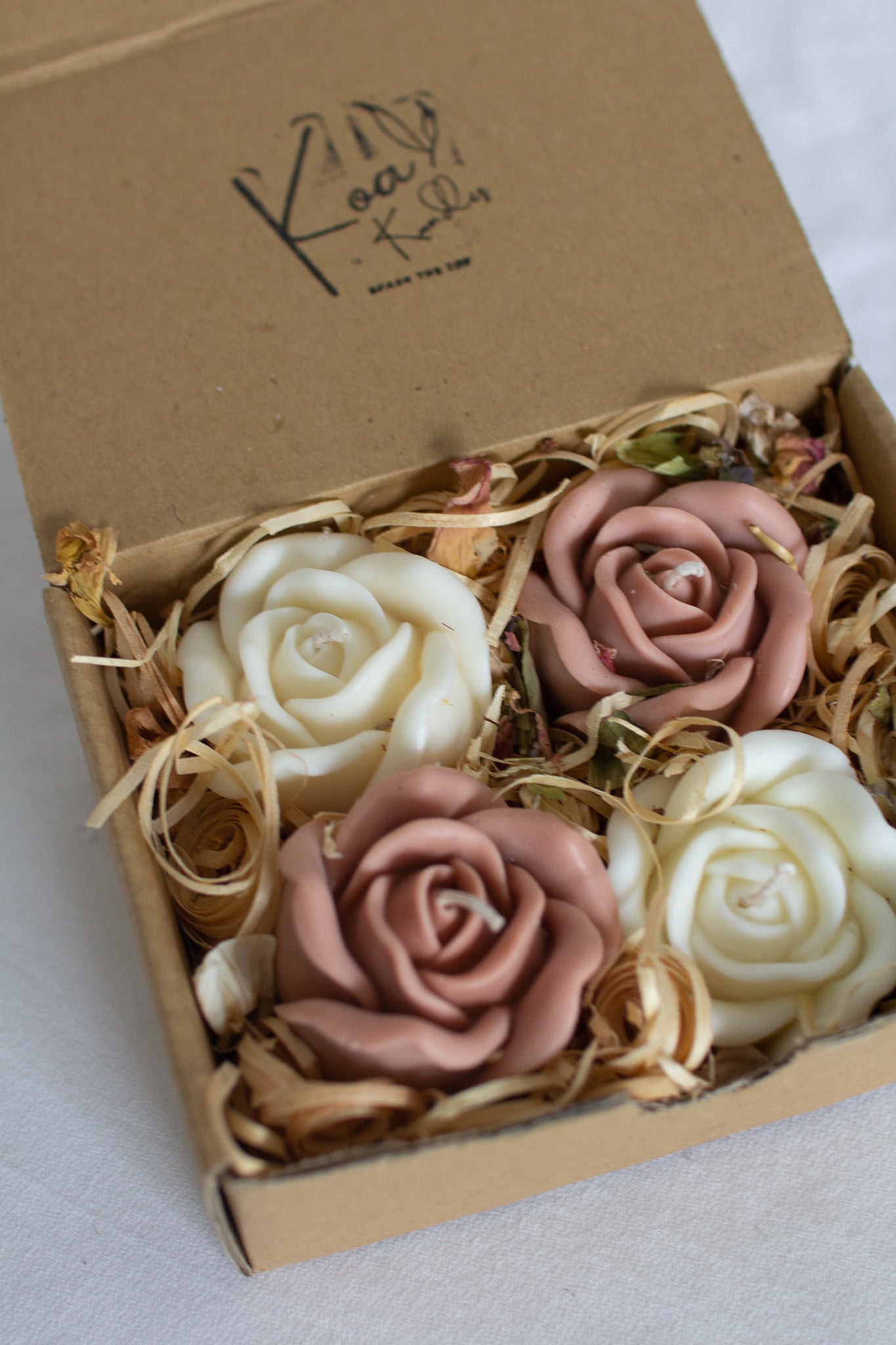 A gift box opened to reveal four rose-shaped candles inside. Two are white and two are a blush pink colour, all wrapped in a eco kraft box